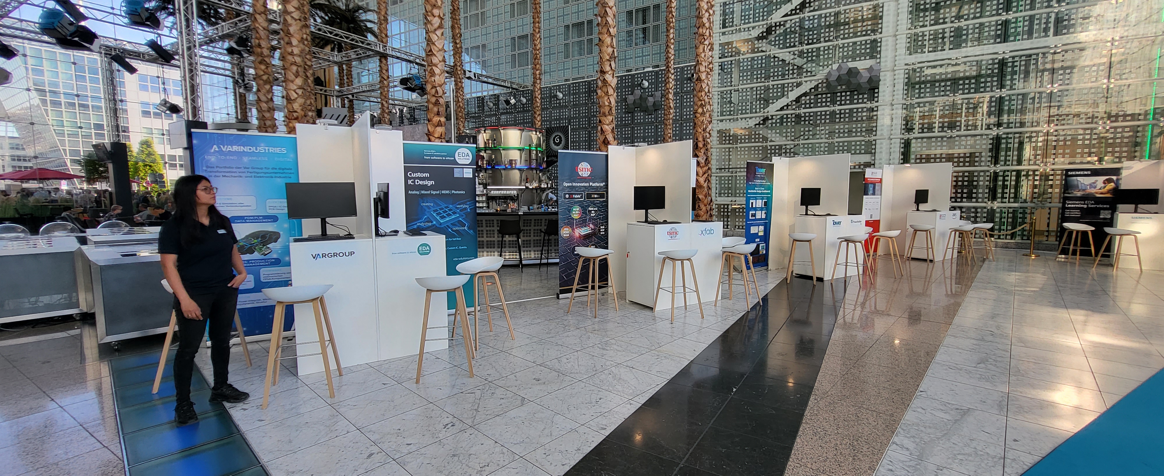 Mehrere Monitore auf Tischen in einer Messehalle mit Palmen